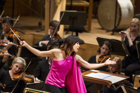 A woman with brown hair in a pink shirt, conductor Sarah Hicks, bows to the audience.