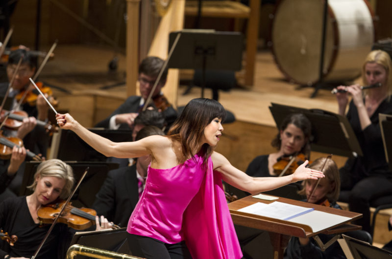 A woman with brown hair in a pink shirt, conductor Sarah Hicks, bows to the audience.
